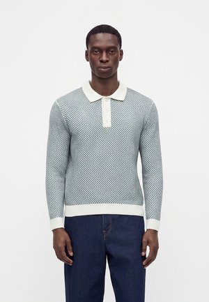 Homme portant un pull polo à manches longues à motifs gris clair et blancs et un jean bleu foncé, debout devant un fond blanc uni.