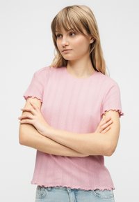 Νέα γυναίκα με ξανθά μαλλιά, φορώντας ροζ πουλόβερ με κοντά μανίκια και μπλε τζιν, στέκεται με τα χέρια σταυρωμένα και κοιτάζει ελαφρώς προς τα πλευρά.