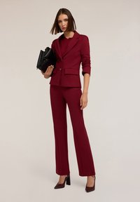 Blazer su misura bordeaux e pantaloni a palazzo coordinati, realizzati in tessuto liscio. La modella tiene in mano una borsa nera ed indossa tacchi a punta.