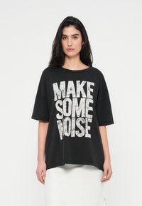 Μαύρο φαρδύ t-shirt με φαρδιά λαιμόκοψη, με μεγάλα λευκά φθαρμένα γράμματα που λένε "ΚΑΝΕ ΛΙΓΟ ΘΟΡΥΒΟ." Κοντά μανίκια και άνετη εφαρμογή.