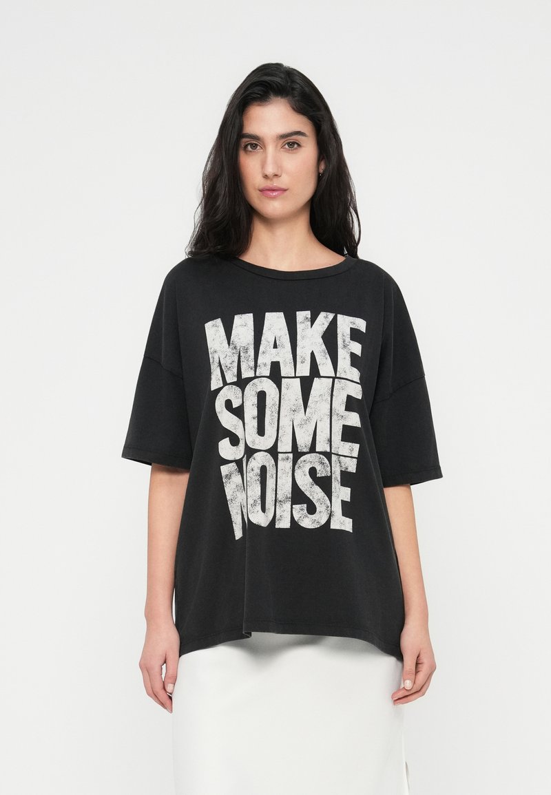Μαύρο φαρδύ t-shirt με φαρδιά λαιμόκοψη, με μεγάλα λευκά φθαρμένα γράμματα που λένε "ΚΑΝΕ ΛΙΓΟ ΘΟΡΥΒΟ." Κοντά μανίκια και άνετη εφαρμογή.