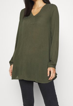 Kvinde iført en olivengrøn tunika med lange ærmer og V-hals samt sorte tætsiddende bukser, stående foran en ensartet baggrund.