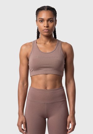 Fit Frau mit geflochtenem Haar, die einen mauvefarbenen Sport-BH und hochgeschnittene Leggings trägt, steht vor einem schlichten hellen Hintergrund.
