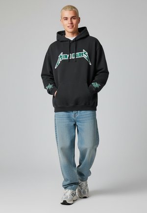 Felpa nera con testo grafico verde e decorazioni a forma di stella, abbinata a jeans blu larghi e sneakers grigie. Texture liscia, design casual.
