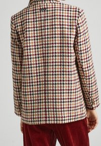 Blazer de lana con un patrón de espiga, que presenta tonos de burdeos, crema, marrón y negro, con botones en las mangas y un corte ajustado.