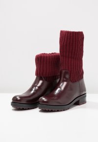 Bottes basses en cuir bordeaux avec poignets en tricot côtelé épais, talons bas et semelles à crampons, présentées sur une surface blanche avec un fond blanc.