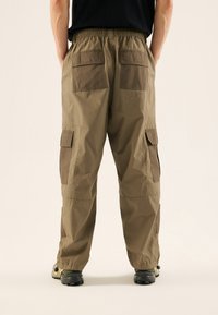Pantalones cargo beige con cinturilla elástica, múltiples bolsillos cargo y paneles marrones en contraste en los lados y la parte trasera. Tejido ligero texturizado.