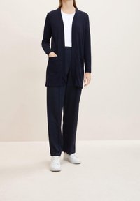 Cardigan blu navy con maniche lunghe e frontale aperto, dotato di due tasche anteriori e una texture liscia, abbinato a pantaloni coordinati e sneakers bianche.