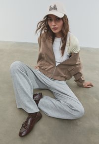 Veste zippée marron sur une chemise blanche, pantalon large gris et chaussures marron à lacets, complété par une casquette beige avec le texte "LA".