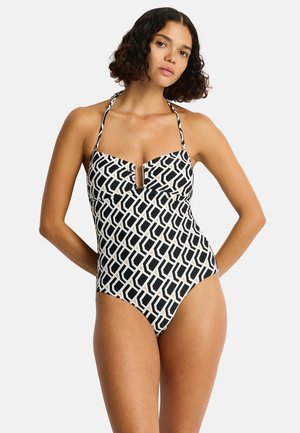 Frau mit lockigem dunklem Haar, die einen einteiligen Badeanzug mit schwarz-weißem geometrischem Muster, Neckholder-Trägern und einem vorderen Ausschnitt trägt.