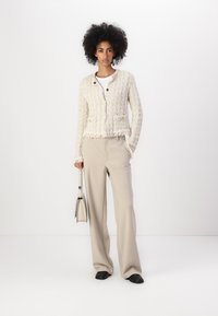 Giacca bianca testurizzata con bordi frangiati, design aderente, due tasche, abbinata a pantaloni beige e con in mano una piccola borsa.