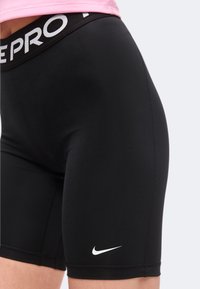 Close-up of black Nike Pro fitted athletic shorts worn with a pink top, showing white Nike swoosh logo on the left thigh.