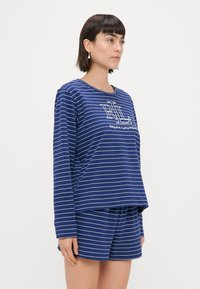 Μπλε και λευκή ριγέ μπλούζα με μακριά μανίκια και στρογγυλή λαιμόκοψη, με το κείμενο "Ralph Lauren." Συνδυάζεται με ριγέ σορτς σε αντίστοιχο χρώμα.