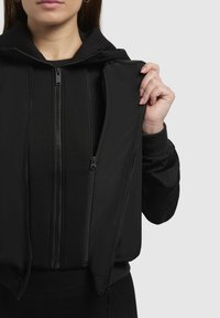 Veste noire à double couche avec un extérieur brillant et un tissu intérieur doux. Présente plusieurs zips et une capuche. Texture lisse avec des manches ajustées.