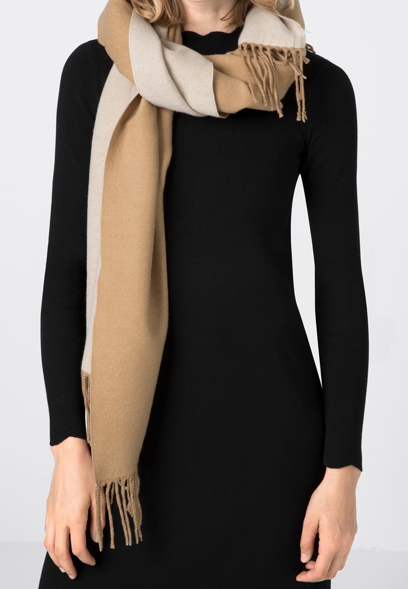 Femme portant une robe noire à manches longues et une épaisse écharpe beige et marron clair avec des franges aux extrémités, enroulée autour de son cou.