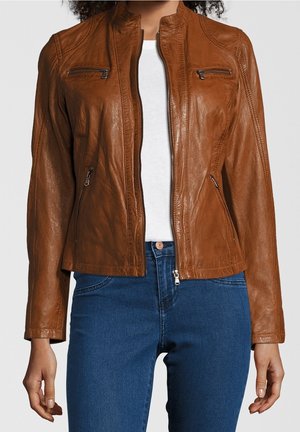 Frau trägt eine eng anliegende braune Lederjacke mit Reißverschlusstaschen über einem weißen Hemd und blauen Jeans, von den Schultern bis zu den Hüften gezeigt.