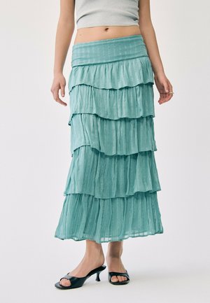 Vue de la taille aux pieds d’une personne portant une jupe longue à volants bleu turquoise clair avec des motifs à rayures verticales et des sandales à talons noires à bout ouvert.