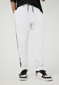 Pantaloni sportivi bianchi con strisce laterali nere, dotati di una vita elastica con cordino e due tasche laterali. Il materiale è morbido e liscio.