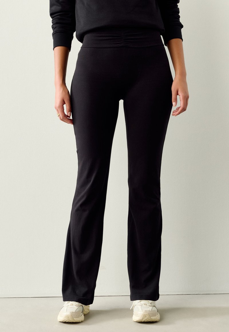 Personne portant un pantalon noir ajusté et des baskets blanches, debout contre un mur clair uni.