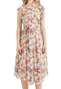 Frau in einem ärmellosen, weißen Midikleid mit orangefarbenen, roten und gelben Blumenmustern sowie einem Taillengürtel.