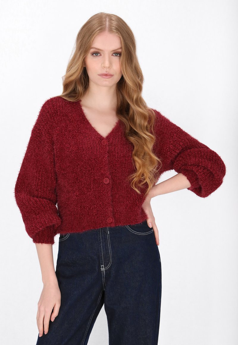 Czerwony sweter dzianinowy z głębokim dekoltem w kształcie V, oversize'owymi rękawami i dwoma guzikami z przodu. Miękka, teksturowana tkanina i krótki krój. Łączony z ciemnymi dżinsami.