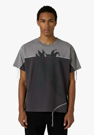 Un giovane indossa una maglietta ampia bicolore grigio e nero con una grafica di cappello da giullare e fili svolazzanti sulle maniche e sull'orlo, su sfondo bianco.