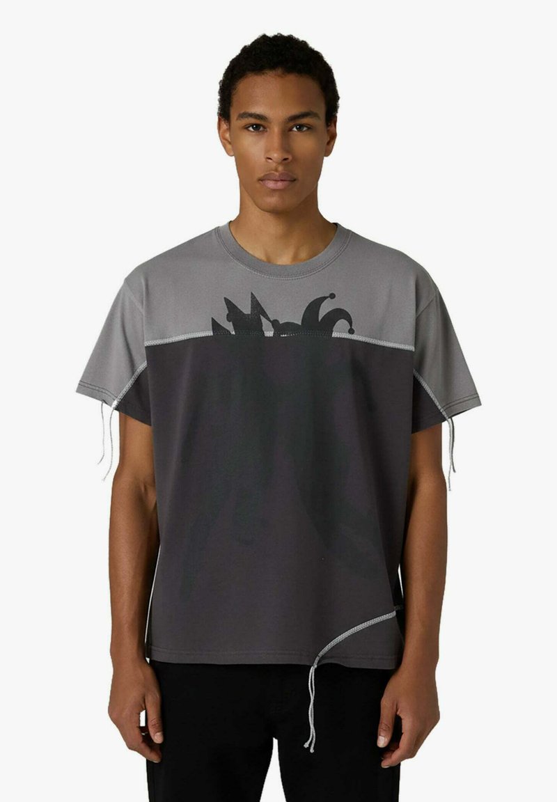 Un giovane indossa una maglietta ampia bicolore grigio e nero con una grafica di cappello da giullare e fili svolazzanti sulle maniche e sull'orlo, su sfondo bianco.