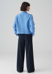 Giacca di lana testurizzata azzurro chiaro con taglio quadrato, maniche lunghe e colletto alto, abbinata a pantaloni neri a gamba larga.