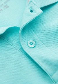 Turkos pikétröja med en texturerad tygkvalitet, med knäppning och krage. Materialet verkar mjukt och andningsbart.