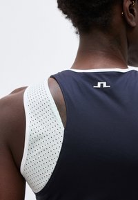 Κοντινό πλάνο ενός ατόμου που φοράει αμάνικο αθλητικό τοπ με σκούρο μπλε πίσω μέρος και λευκή διάτρητη πλευρική επιφάνεια, με ένα μικρό λογότυπο κάτω από τη γραμμή του λαιμού.