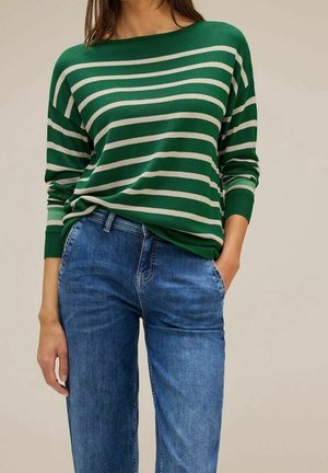 Vrouw draagt een groen-wit gestreept shirt met lange mouwen in combinatie met blauwe spijkerbroek, met één hand in de zak tegenover een effen achtergrond.