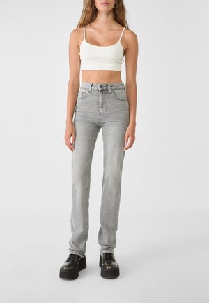 Femme portant un débardeur court blanc, un jean droit taille haute gris clair et des chaussures à plateforme noires à lacets, debout sur un fond blanc.