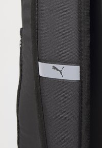 Sac à dos noir avec une surface texturée, comprenant un panneau gris avec un logo imprimé. Inclut des zips latéraux et des coutures renforcées.