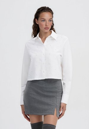 Giovane donna con capelli mossi indossa una camicia bianca texturizzata con bottoni e una mini gonna grigia a righe sottili con uno spacco laterale.