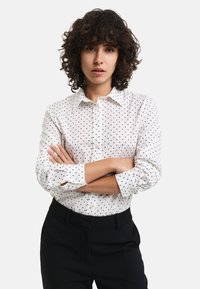 White button-up shirt with black polka dots, featuring a classic collar and long sleeves. Paired with black trousers.