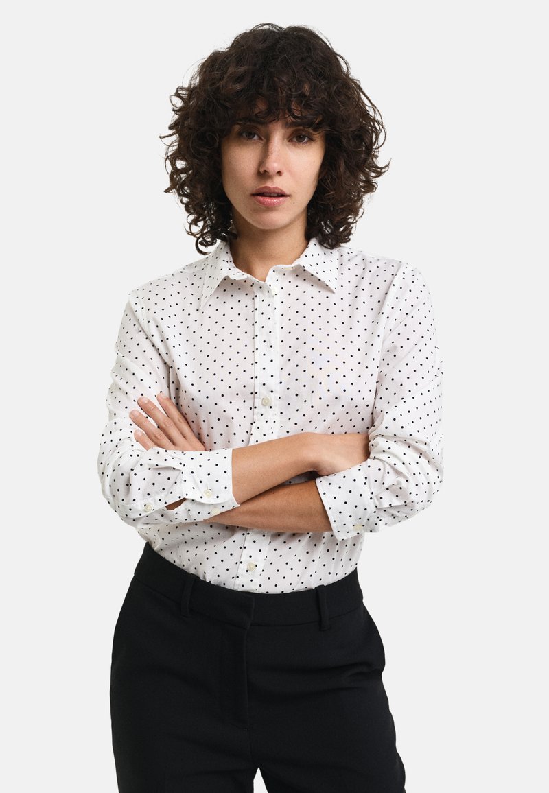 White button-up shirt with black polka dots, featuring a classic collar and long sleeves. Paired with black trousers.
