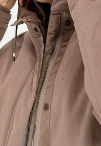Chaqueta marrón con cierre de cremallera y botones en la parte delantera, con textura acolchada, capucha ajustable y detalle de tirador de cremallera en contraste.