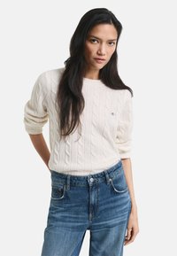 Maglione crema a treccia con collo rotondo e maniche arrotolate, abbinato a jeans blu in denim a vita alta con chiusura a bottone.