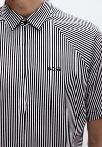 Cămașă cu dungi negre și albe cu mânecă scurtă, cu guler clasic; are un mic logo negru pe piept. Textură de bumbac.