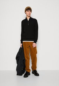 Black zip-up sweater, brown corduroy pants, black shoes, and dark jacket held. Warm knit hat complements the outfit's color scheme.