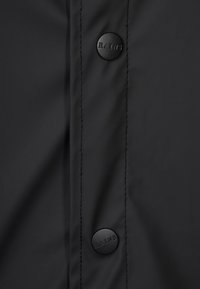 Mustan vedenpitävän takin lähikuva, jossa näkyy sileä pinta, ompeleet ja kaksi mustaa painonappia, joissa on kohokuvioitu "RAINS".