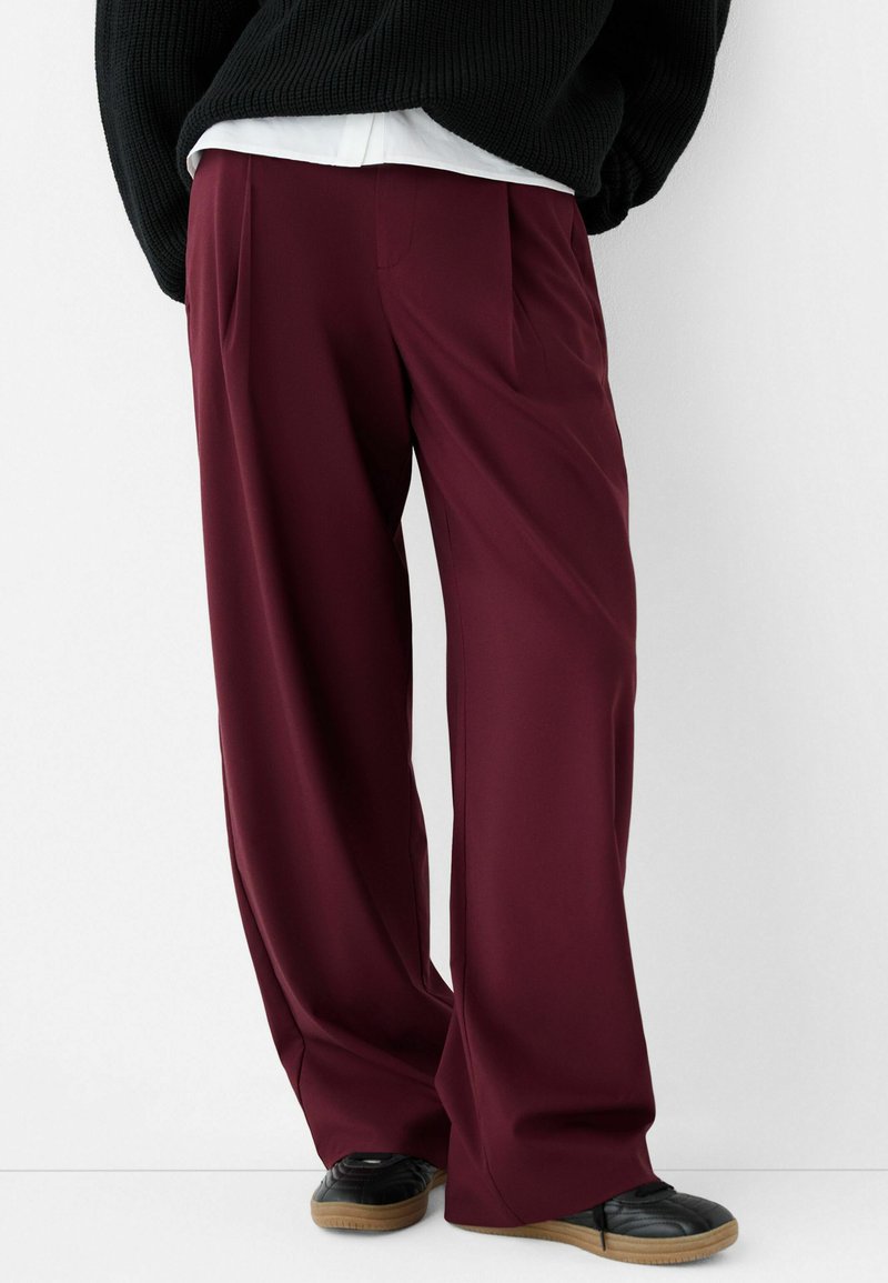 Pantalons larges bordeaux avec plis avant, confectionnés en tissu lisse, associés à des chaussures noires avec une tige texturée et une semelle en gomme.