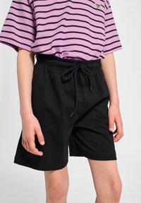 Schwarze Baumwollshorts mit elastischem Bund und Kordelzug, kombiniert mit einem lila-schwarz gestreiften kurzärmeligen Hemd.