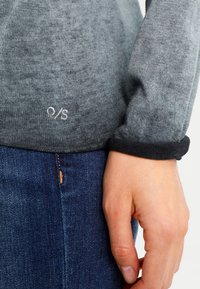 Grå sweater med svarta ärmslut, textilstruktur och liten vit "O/S" logotyp vid fållen. Bärs med blå denimjeans.