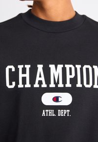 Black sweatshirt featuring "CHAMPION" in bold white letters and "ATHL. DEPT." below. Includes a logo in red and blue within an oval.