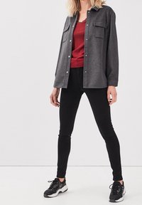 Szary koszula z guzikami z dwiema kieszeniami na piersi, nałożona na czerwoną bluzkę, w zestawie z czarnymi obcisłymi dżinsami i czarnymi trampkami. Białe tło.