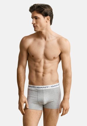 Muscular man with short dark hair wearing grey GANT boxer briefs, standing against plain light background, looking to his left.