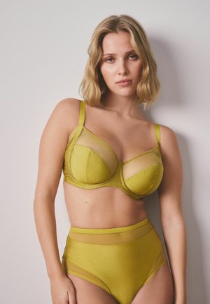 Frau mit schulterlangem blondem Haar, die einen gelb-grünen transparenten Mesh-BH und hoch taillierte Höschen trägt, vor einer schlichten hellen Wand stehend.