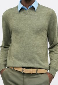 Homme portant une chemise bleu clair à col sous un pull vert olive à col en V avec un pantalon olive assorti et une ceinture beige, mains dans les poches.