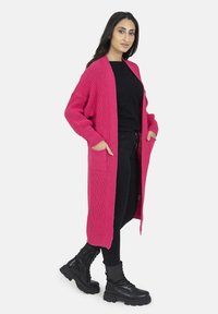 Lång rosa kofta i strukturerad stickad tyg, med fickor och öppen framsida, ihop med en svart topp och byxor. Svarta kängor.